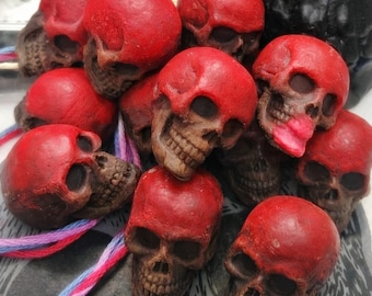 May include: A collection of small, molded skull figurines featuring a vibrant red finish on the cranium and a dark brown, weathered texture on the lower facial structure. One skull displays a bright pink accent on the lower jaw area.