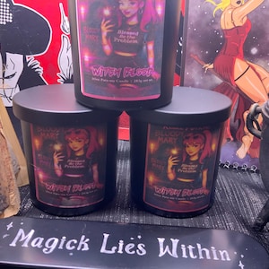 May include: Three black candles with labels featuring a cartoon woman with pink hair and the text "Witch Blood". The candles are stacked, with the text "Magick Lies Within" on a black oval in the foreground. A cauldron is visible.