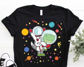 May include: Black t-shirt with a cartoon illustration of a dinosaur astronaut holding a smaller dinosaur in a space helmet. The design includes planets, stars, and a comet.