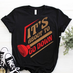 Peut inclure: T-shirt noir avec un graphique brun et rouge qui dit "It's about to go down" avec une illustration de piston.