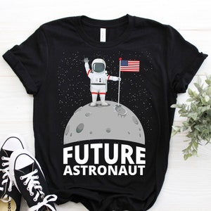 May include: Black t-shirt featuring a cartoon astronaut on the moon holding an American flag. The words "FUTURE ASTRONAUT" are printed in white below the moon. Black and white sneakers are in the foreground.