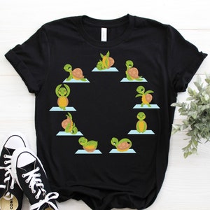 Op de afbeelding: Zwart T-shirt met een afbeelding van schildpadden die yoga-houdingen aannemen op blauwe matten. De schildpadden zijn groen met bruine schilden en zijn in een cirkel gerangschikt.