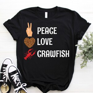 May include: Black t-shirt with the words "PEACE LOVE CRAWFISH" in white. The design includes a peace sign, a leopard print heart, and a red crawfish. Black and white sneakers are in the foreground.