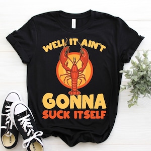 May include: Black t-shirt with the text "WELL IT AIN'T GONNA SUCK ITSELF" in yellow and orange. A red crawfish is centered above the text. Black and white trainers are in the foreground.