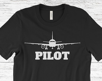 May include: Black t-shirt featuring a white graphic of an aeroplane and the word "PILOT" in capital letters. The aeroplane is positioned above the text, with its wings and tail visible. The shirt is made of a soft material.