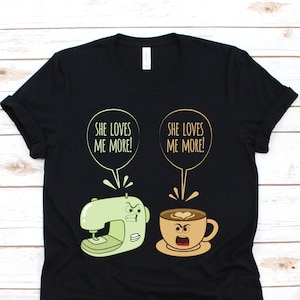 May include: Black t-shirt with a humorous graphic featuring a sewing machine and a coffee cup, each with an angry expression. Text above the images reads "SHE LOVES ME MORE!" in a speech bubble.