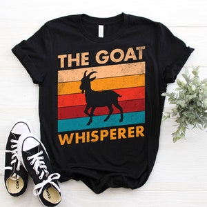 Puede incluir: Camiseta negra con el texto "THE GOAT WHISPERER" encima y debajo de una silueta de cabra. El texto y la cabra est&aacute;n sobre un fondo retro de rayas naranjas, amarillas, rojas y turquesas. Zapatillas de lona negras con cordones blancos en primer plano.
