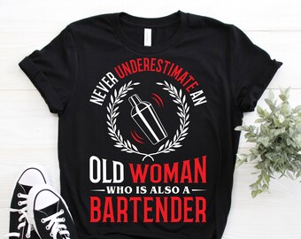May include: Black t-shirt with white text that reads "Never underestimate an old woman who is also a bartender". The text is surrounded by a white wreath with a cocktail shaker in the center.