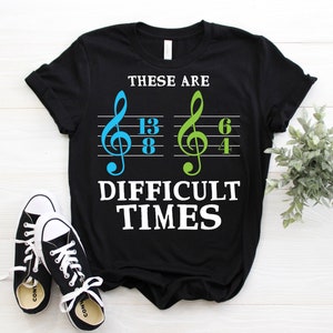 Könnte beinhalten: Schwarzes T-Shirt mit dem Text "THESE ARE DIFFICULT TIMES" und Noten. Die Noten sind blau und grün. Ein Paar schwarz-weiße Turnschuhe sind im Vordergrund.