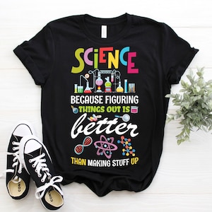 May include: Black t-shirt with the word "SCIENCE" in colorful letters, featuring laboratory equipment graphics. The text "BECAUSE FIGURING THINGS OUT IS better THAN MAKING STUFF UP" is also displayed. Black and white sneakers are in the foreground.