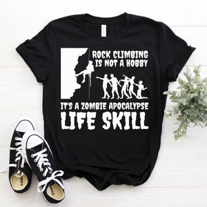 Puede incluir: Camiseta negra con texto gráfico e ilustraciones en blanco. El texto dice: "Escalar no es un hobby, es una habilidad para la vida en un apocalipsis zombi". Incluye un escalador y figuras de zombis. Zapatillas negras y blancas en primer plano.