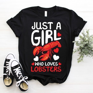 May include: Black t-shirt with the text "JUST A GIRL WHO LOVES LOBSTERS" in white and red. The design features a red lobster and red hearts. A pair of black and white trainers are in the foreground.