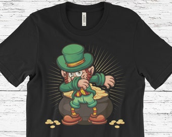 Puede incluir: Camiseta negra con un dibujo de un duende haciendo un dab junto a una olla de oro. El duende lleva un sombrero, chaqueta y pantalones verdes, con zapatos naranjas. Monedas de oro están esparcidas alrededor de la olla.