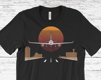 May include: Black t-shirt with a graphic of an aeroplane landing on a runway, set against a sunset. The design includes a stylized aeroplane, airport buildings, and a gradient sunset in shades of orange and red.