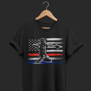 May include: Black t-shirt with a white silhouette of a baseball player catching a ball in front of an American flag with a red stripe and a blue stripe.