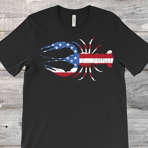 May include: Black t-shirt with a distressed crawfish graphic. The crawfish design incorporates the American flag's stars and stripes in red, white, and blue. The shirt is made of a soft material.
