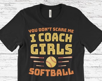 May include: Black t-shirt with the text "YOU DON'T SCARE ME I COACH GIRLS SOFTBALL". The design includes a softball, bats, and text in orange and yellow. A casual tee for softball coaches.