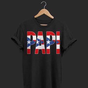 May include: Black t-shirt with the word "PAPI" in large, bold letters. The letters are designed with the Puerto Rican flag's colors: red, white, and blue, with stars. The shirt is displayed on a wooden hanger.