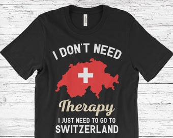 Può includere: T-shirt nero con la scritta "I DON'T NEED Therapy I JUST NEED TO GO TO SWITZERLAND." Il design include un contorno rosso della Svizzera con una croce bianca al centro.