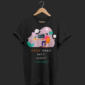 May include: Black t-shirt with a graphic of a person coding at a desk, with the text "while !dead: eat() code() //sleep()" in orange and green. The design includes a computer, a clock, and a speech bubble with code symbols.
