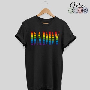Op de afbeelding: Zwart t-shirt met het woord "DADDY" in regenboogkleuren. De tekst is ontworpen met een golvend, blokkerig lettertype. Het shirt hangt aan een houten hanger.