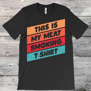 Puede incluir: Camiseta negra con el texto "THIS IS MY MEAT SMOKING T-SHIRT" en rayas diagonales naranjas, rojas y azules. La camiseta está hecha de un material suave y es adecuada para uso casual.