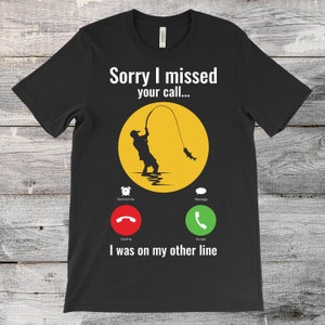 May include: Black t-shirt with a fishing graphic. The design includes a silhouette of a person fishing, a yellow circle, and the text "Sorry I missed your call..." with phone interface icons and the text "I was on my other line."