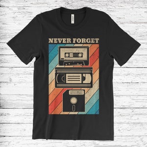 May include: Black t-shirt with a retro-style graphic of a cassette tape, VHS tape, and floppy disk. The graphic is layered on a rainbow background with the text "NEVER FORGET" above it.