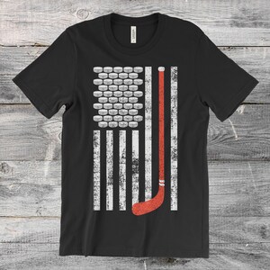 May include: Black t-shirt featuring a distressed graphic of the American flag, with hockey pucks replacing the stars and a hockey stick in place of a stripe. The design is in white and red.
