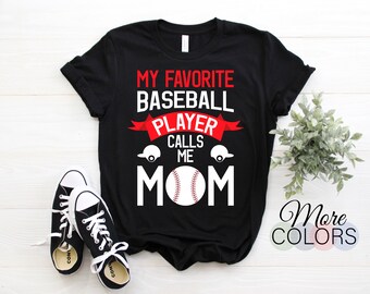 May include: Black t-shirt with the text "MY FAVORITE BASEBALL PLAYER CALLS ME MOM" in red and white. The design includes a baseball, baseball caps, and a red banner. A pair of black and white sneakers are in the foreground.