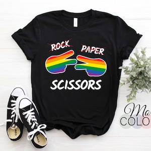 Pode incluir: Camiseta preta com uma mão arco-íris fazendo o sinal de tesoura. As palavras "Pedra Papel Tesoura" estão impressas na camiseta, com "Pedra" e "Papel" riscadas.
