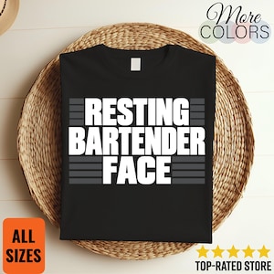 May include: A black short-sleeved t-shirt featuring the white text "RESTING BARTENDER FACE" centred on the chest. The text is surrounded by horizontal grey lines above and below the words, creating a bold graphic design on the front of the shirt.