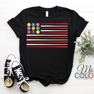 May include: Black t-shirt featuring a graphic design resembling the American flag, with billiard balls replacing the stars and stripes. The stripes alternate between red and white. The shirt is paired with black and white sneakers. The text "More Colors" is visible.