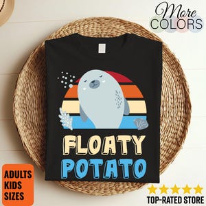 Puede incluir: Camiseta negra con un diseño de foca de dibujos animados y las palabras "FLOATY POTATO" en amarillo y azul. El diseño incluye un fondo de puesta de sol y el texto "More Colors". La camiseta se muestra en una cesta tejida.