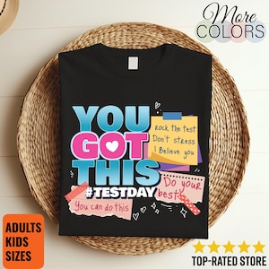 Könnte beinhalten: Schwarzes T-Shirt mit dem Text "YOU GOT THIS" in Blau und Pink. Zusätzlicher Text enthält "#TESTDAY", "You can do this", "Rock the test, Don't stress, I Believe you" und "Do your best". Das Shirt wird auf einem geflochtenen Korb präsentiert.