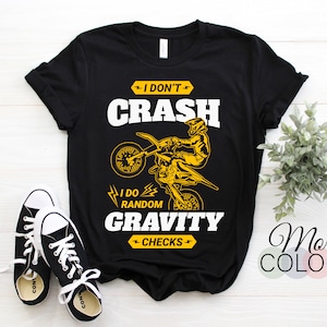 以下が含まれることがあります： 黒いTシャツ。黄色のモトクロスライダーのグラフィックと「I DON'T CRASH I DO RANDOM GRAVITY CHECKS」の文字。シャツは黒と白のスニーカーと組み合わせて表示されます。「More COLORS」の文字も表示されます。