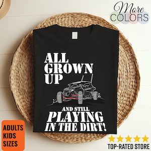 Może przedstawiać: Czarny t-shirt z białą grafiką pojazdu terenowego oraz napisem ALL GROWN UP AND STILL PLAYING IN THE DIRT. Projekt zawiera czerwone akcenty na zawieszeniu pojazdu oraz białe ślady opon na podłożu znajdującym się pod samym pojazdem.