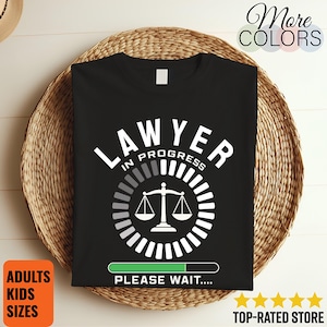 May include: A black t-shirt featuring white text that reads LAWYER IN PROGRESS. Below the text is a circular loading icon with a scale of justice in the center. At the bottom, a green progress bar is displayed with the text PLEASE WAIT....