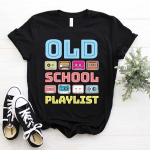 May include: Black t-shirt with the words "OLD SCHOOL PLAYLIST" in a retro font. The design includes colorful cassette tape illustrations. A pair of black and white sneakers are in the foreground.