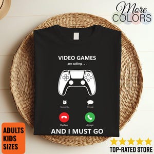 Op de afbeelding: Een zwart t-shirt met een witte afbeelding van een videogamecontroller. De tekst "VIDEO GAMES are calling..." staat boven de controller en "AND I MUST GO" eronder, met rode en groene oproeppictogrammen in het midden.