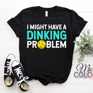 Peut inclure: T-shirt noir avec le texte "I might have a dinking problem" en blanc avec un "dinking" turquoise et un graphique de pickleball jaune.