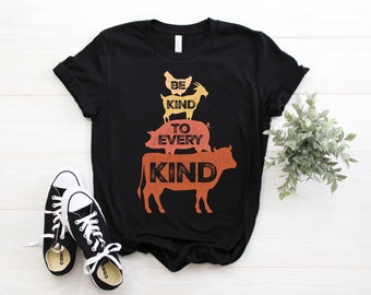 Könnte beinhalten: Schwarzes T-Shirt mit einem Grafikdruck einer Kuh, eines Schweins, einer Ziege und eines Huhns, die übereinander gestapelt sind. Der Text "Be Kind To Every Kind" ist auf dem Shirt aufgedruckt.