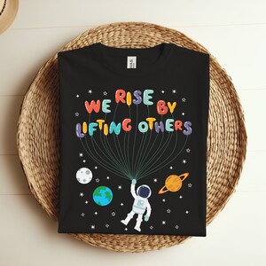 Pode incluir: Camiseta preta com um gráfico de balões coloridos que soletram "WE RISE BY LIFTING OTHERS" e um astronauta segurando os balões. O fundo é um espaço preto com estrelas e planetas.