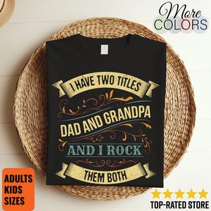 Pode incluir: Camiseta preta com o texto "I HAVE TWO TITLES DAD AND GRANDPA AND I ROCK THEM BOTH" em um design decorativo. A camisa é exibida em uma cesta de vime. O texto adicional inclui "ADULTS KIDS SIZES" e "TOP-RATED STORE".