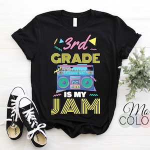 Puede incluir: Camiseta negra con un gráfico retro de boombox y el texto "3rd Grade Is My Jam". El diseño presenta acentos rosas, amarillos y azules. Un par de zapatillas negras y blancas están en primer plano.