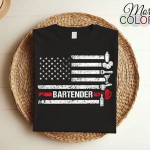 Pode incluir: Camiseta preta com um design de bandeira americana desbotada e a palavra "BARTENDER" em vermelho. A bandeira tem um campo de estrelas branco e listras vermelhas e brancas. Há um coqueteleira branca, uma taça de martini e outras ferramentas de bar na bandeira.