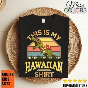 May include: Black t-shirt with a graphic of a dinosaur wearing a Hawaiian shirt, with the text "THIS IS MY HAWAIIAN SHIRT". The shirt has a retro sunset design and is on a woven basket.