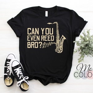 Op de afbeelding: Zwart T-shirt met een gouden afbeelding van een saxofoon en de tekst "Can you even reed bro?"
