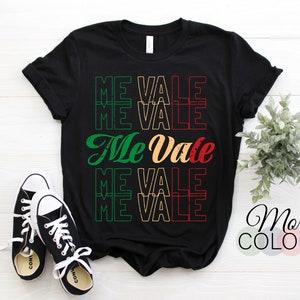 May include: Black t-shirt with the phrase "Me Vale" repeated in green, red, and yellow. The text has a distressed, vintage look. The shirt is laid flat on a white surface with a pair of black sneakers and a small potted plant.