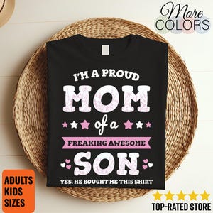 Könnte beinhalten: Schwarzes T-Shirt mit dem Text "I'M A PROUD MOM of a FREAKING AWESOME SON". Das Wort "MOM" hat ein florales Design. Pinke Sterne und Herzen akzentuieren den Text. Das Shirt wird auf einem geflochtenen Korb präsentiert.
